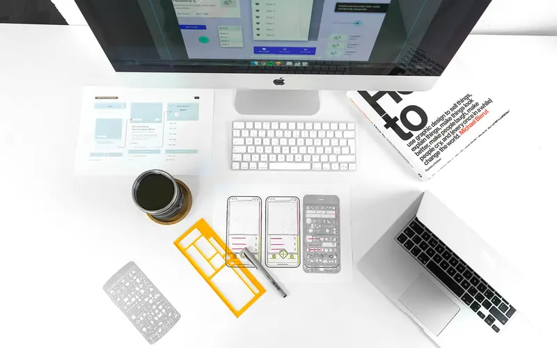 An overhead view of a desk with a keyboard, a laptop, phone prototypes and printed screen layouts